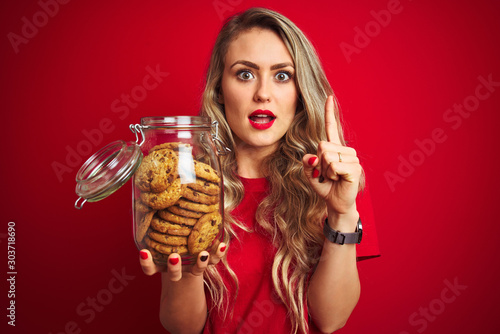 Young beautiful woman holding a jar of cookies over red isolated background surprised with an idea or question pointing finger with happy face, number one