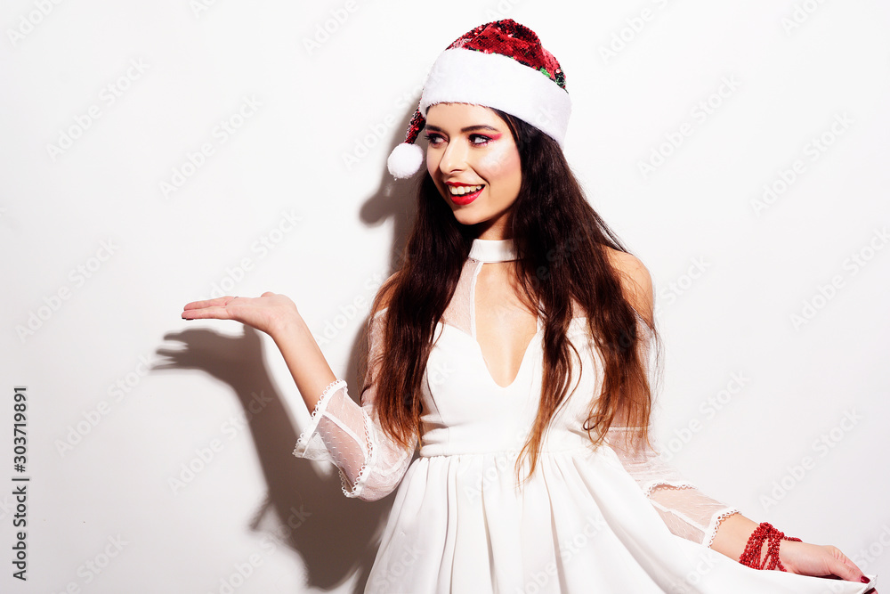 Beautiful brunette girl in santa hat shows to the side, holds your product, free space for text Beautiful brunette girl in santa hat shows to the side, holds your product, free space for text