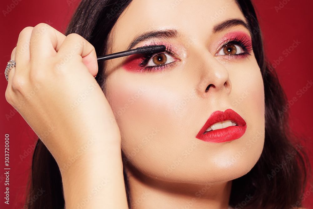 Portrait, model holds mascara in hands and paints eyelashes. Beautiful brunette with professional red make-up, on a red background. New Year's red make-up