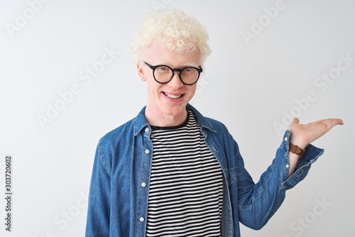 Young albino blond man wearing denim shirt and glasses over isolated white background smiling cheerful presenting and pointing with palm of hand looking at the camera.