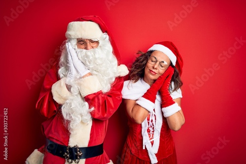 Middle age couple wearing Santa costume and glasses over isolated red background sleeping tired dreaming and posing with hands together while smiling with closed eyes.
