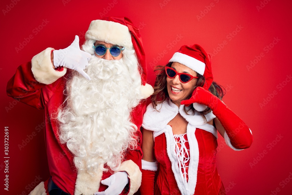 Middle age couple wearing Santa costume and sunglasses over isolated red background smiling doing phone gesture with hand and fingers like talking on the telephone. Communicating concepts. Middle age couple wearing Santa costume and sunglasses over isolated red background smiling doing phone gesture with hand and fingers like talking on the telephone. Communicating concepts.