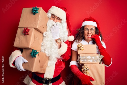 Middle age couple wearing Santa costume holding tower of gifts over isolated red background looking stressed and nervous with hands on mouth biting nails. Anxiety problem.