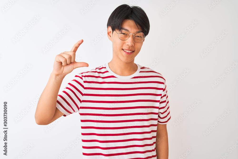 Young chinese man wearing glasses and striped t-shirt standing over isolated white background smiling and confident gesturing with hand doing small size sign with fingers looking and the camera. Young chinese man wearing glasses and striped t-shirt standing over isolated white background smiling and confident gesturing with hand doing small size sign with fingers looking and the camera.
