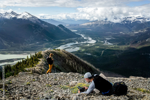 Young adventure photographer taking picture of woman model in yellow trousers and brown hat hiking Greenock trail in Jasper National park, Canada. Athabasca river. Taking photos for social media