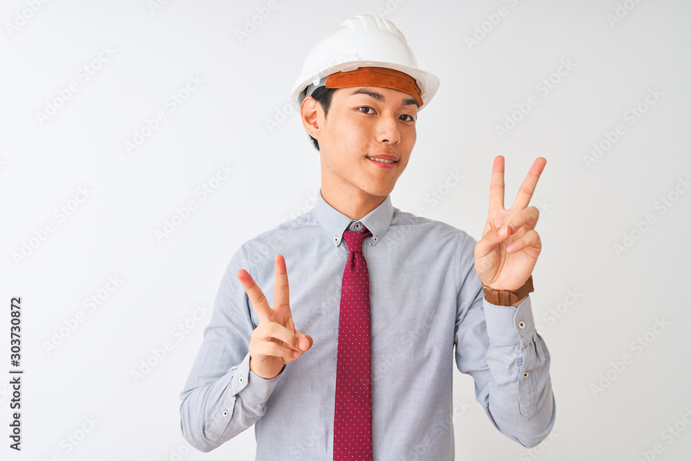 Chinese architect man wearing tie and helmet standing over isolated white background smiling looking to the camera showing fingers doing victory sign. Number two. Chinese architect man wearing tie and helmet standing over isolated white background smiling looking to the camera showing fingers doing victory sign. Number two.