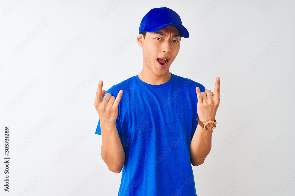 Chinese deliveryman wearing blue t-shirt and cap standing over isolated white background shouting with crazy expression doing rock symbol with hands up. Music star. Heavy concept.