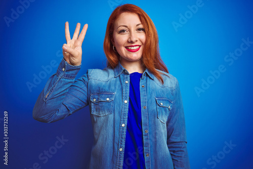 Fotografie, Tablou Young beautiful redhead woman wearing denim shirt standing over blue isolated background showing and pointing up with fingers number three while smiling confident and happy