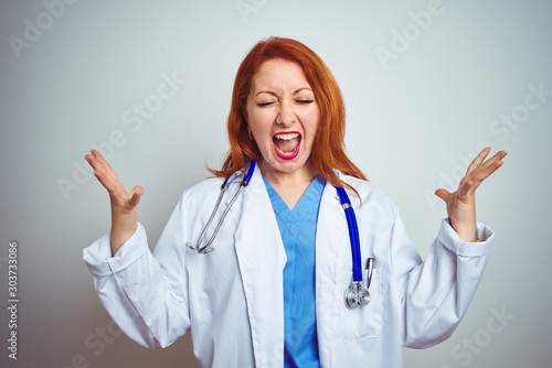 Young redhead doctor woman using stethoscope over white isolated background celebrating mad and crazy for success with arms raised and closed eyes screaming excited. Winner concept