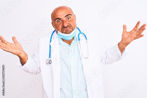 Middle age doctor man wearing stethoscope and mask over isolated white background clueless and confused expression with arms and hands raised. Doubt concept.