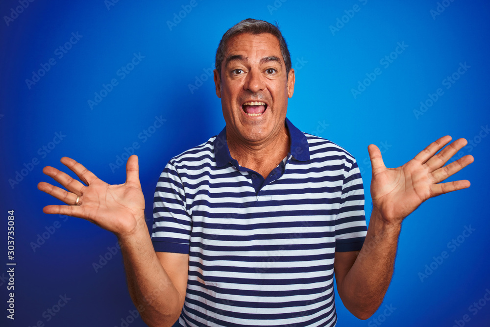 Naklejka premium Handsome middle age man wearing striped polo standing over isolated blue background celebrating crazy and amazed for success with arms raised and open eyes screaming excited. Winner concept