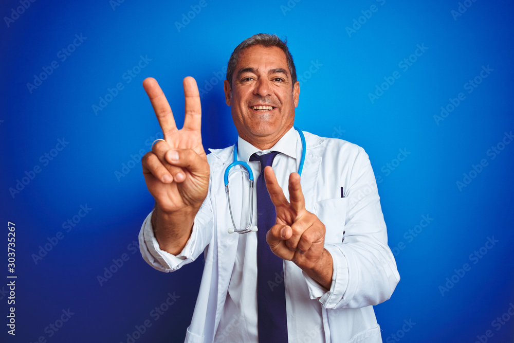 Handsome middle age doctor man wearing stethoscope over isolated blue background smiling looking to the camera showing fingers doing victory sign. Number two. Handsome middle age doctor man wearing stethoscope over isolated blue background smiling looking to the camera showing fingers doing victory sign. Number two.