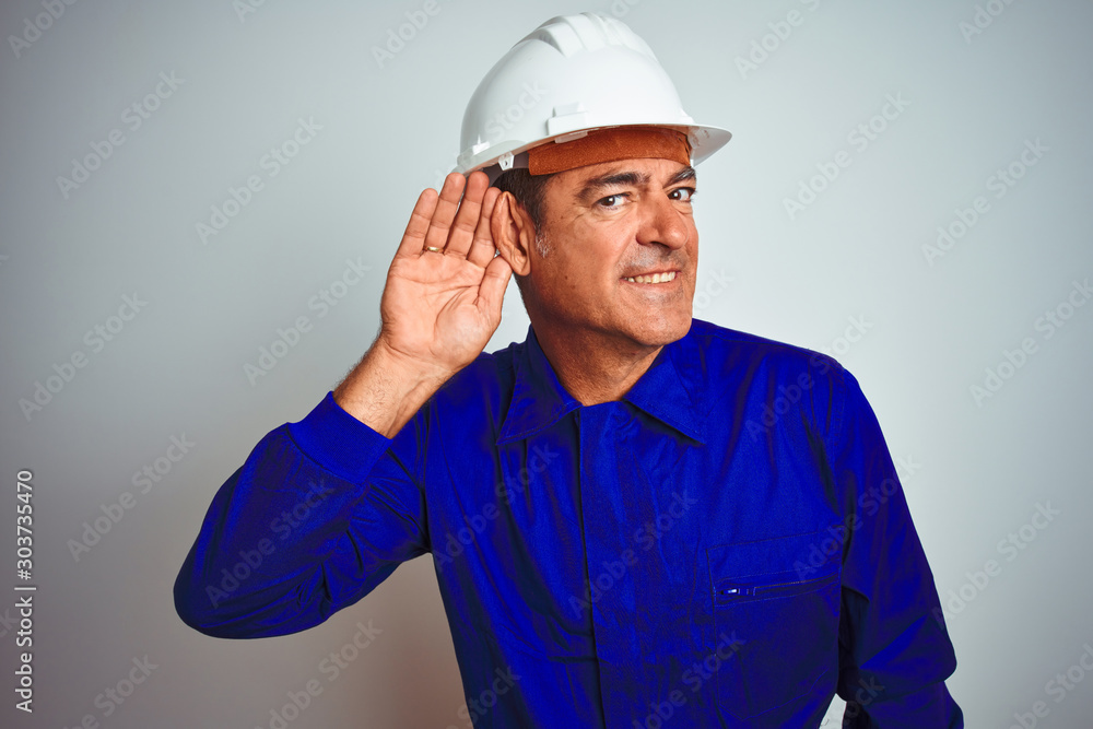 Handsome middle age worker man wearing uniform and helmet over isolated white background smiling with hand over ear listening an hearing to rumor or gossip. Deafness concept. Handsome middle age worker man wearing uniform and helmet over isolated white background smiling with hand over ear listening an hearing to rumor or gossip. Deafness concept.