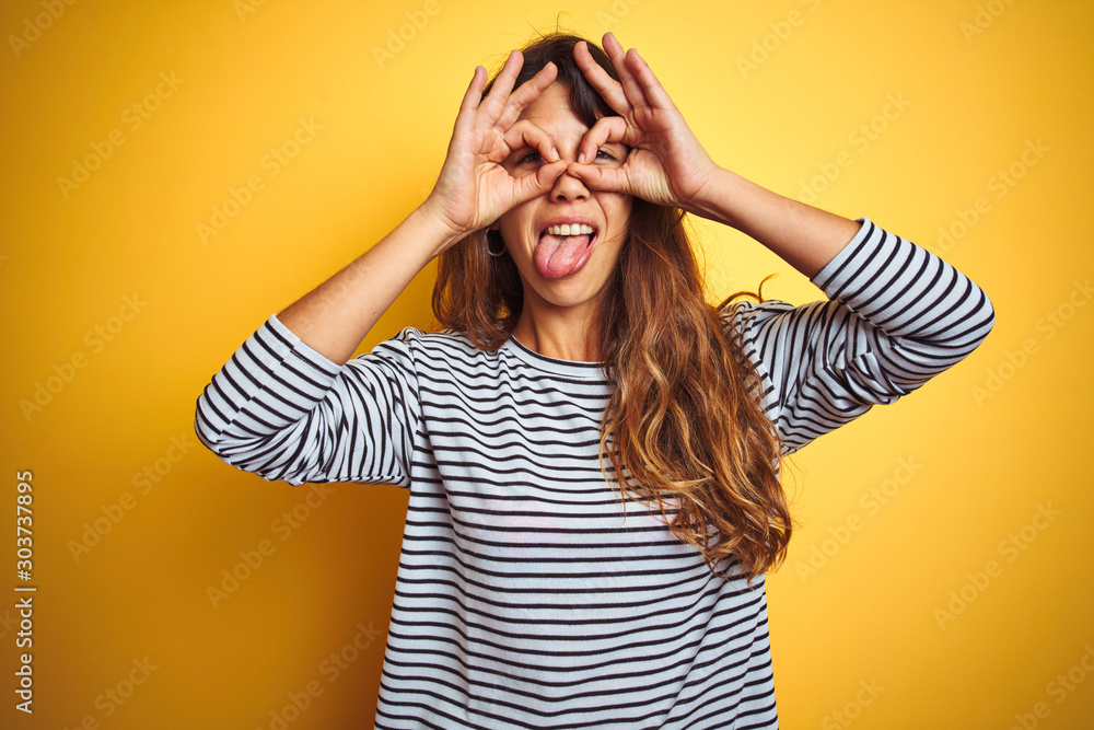 Young beautiful woman wearing stripes t-shirt standing over yelllow isolated background doing ok gesture like binoculars sticking tongue out, eyes looking through fingers. Crazy expression. Young beautiful woman wearing stripes t-shirt standing over yelllow isolated background doing ok gesture like binoculars sticking tongue out, eyes looking through fingers. Crazy expression.