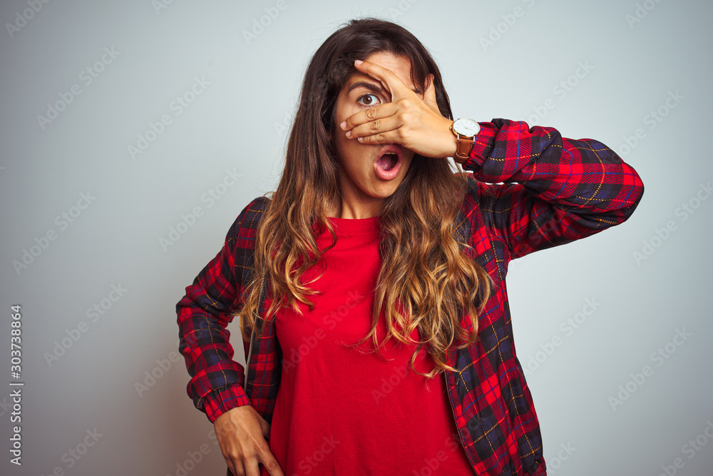 Φωτογραφία Young beautiful woman wearing red t-shirt and jacket standing over white isolated background peeking in shock covering face and eyes with hand, looking through fingers with embarrassed expression