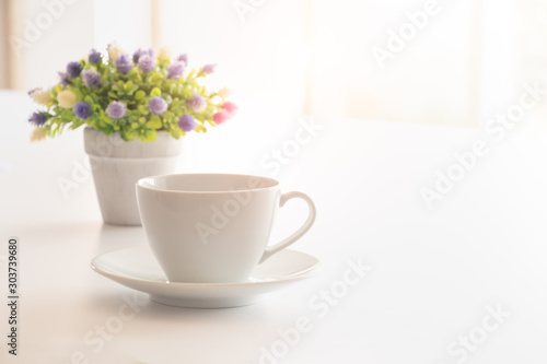 A cup of coffee on white office desk with lovely vase of small flower with morning light flare. A good starting in the early morning to get ready for a day.Free copy space on right for text or design