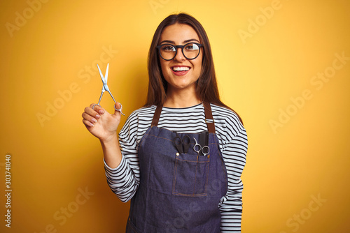 Young beautiful hairdresser woman holding scissors standing over isolated yellow background with a happy face standing and smiling with a confident smile showing teeth photo