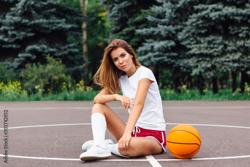 Beautiful young girl dressed in white t-shirt, shorts and sneakers, sits on a basketball court with ball. © Smile