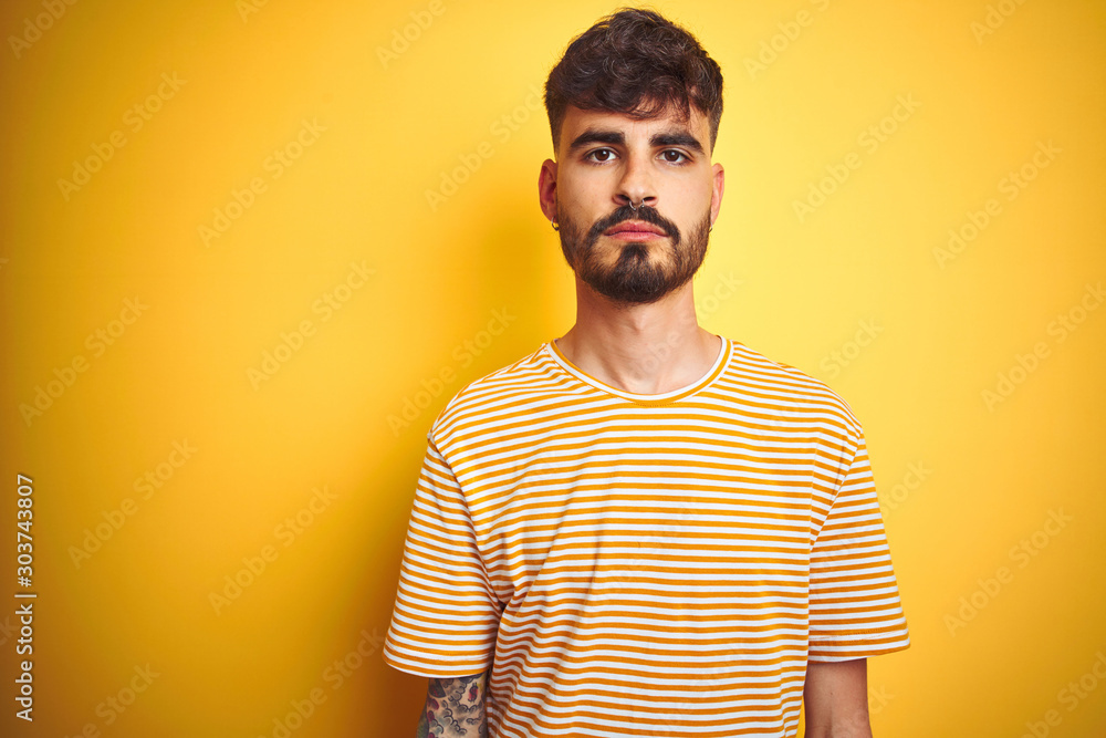 Young man with tattoo wearing striped t-shirt standing over isolated yellow background Relaxed with serious expression on face. Simple and natural looking at the camera.