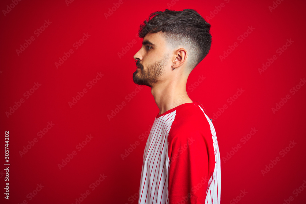 Young man with tattoo wearing striped t-shirt standing over isolated red background looking to side, relax profile pose with natural face with confident smile. Young man with tattoo wearing striped t-shirt standing over isolated red background looking to side, relax profile pose with natural face with confident smile.