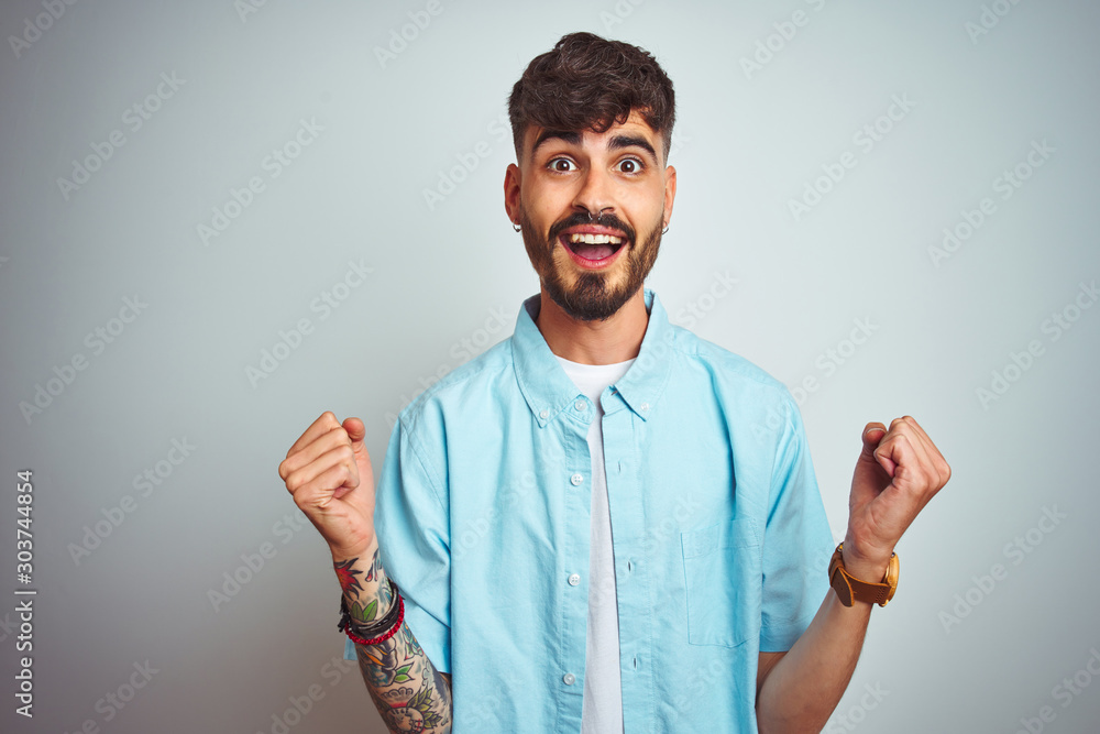 Young man with tattoo wearing blue shirt standing over isolated white background celebrating surprised and amazed for success with arms raised and open eyes. Winner concept. Young man with tattoo wearing blue shirt standing over isolated white background celebrating surprised and amazed for success with arms raised and open eyes. Winner concept.