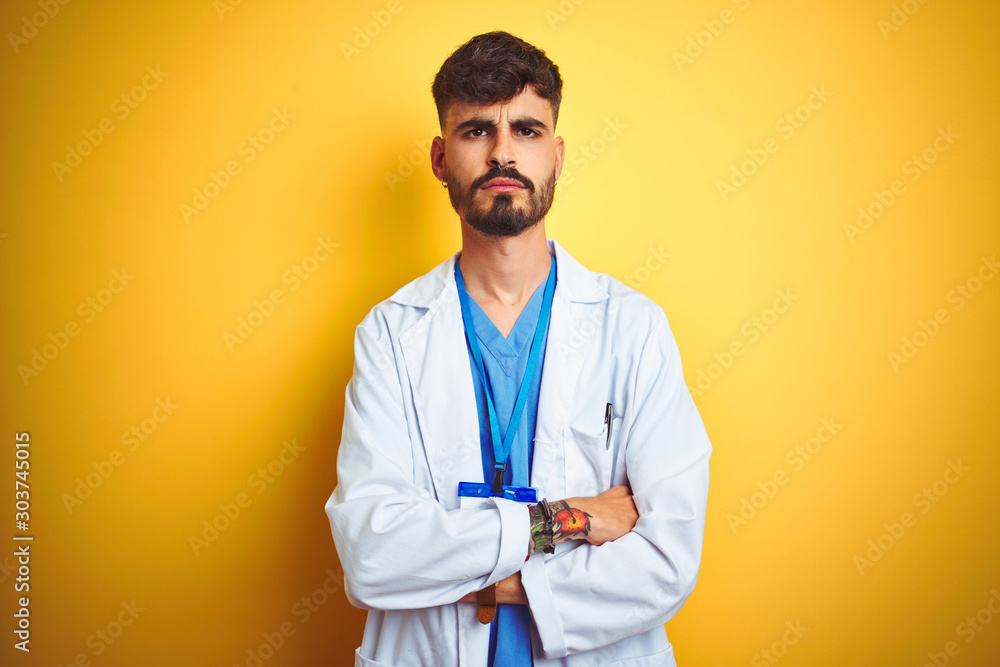 Young doctor man with tattoo wearing id card standing over isolated yellow background skeptic and nervous, disapproving expression on face with crossed arms. Negative person. Young doctor man with tattoo wearing id card standing over isolated yellow background skeptic and nervous, disapproving expression on face with crossed arms. Negative person.