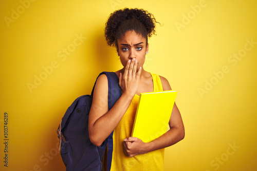 American student woman wearing backpack holding notebook over isolated yellow background cover mouth with hand shocked with shame for mistake, expression of fear, scared in silence, secret concept