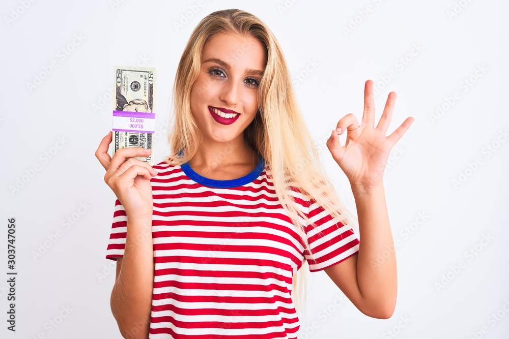 Obraz premium Beautiful woman wearing red striped t-shirt holding dollars over isolated white background doing ok sign with fingers, excellent symbol