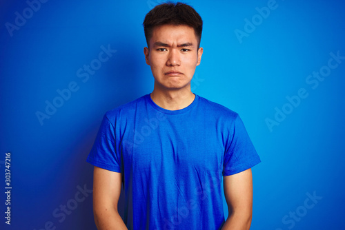 Young asian chinese man wearing t-shirt standing over isolated blue background depressed and worry for distress, crying angry and afraid. Sad expression.