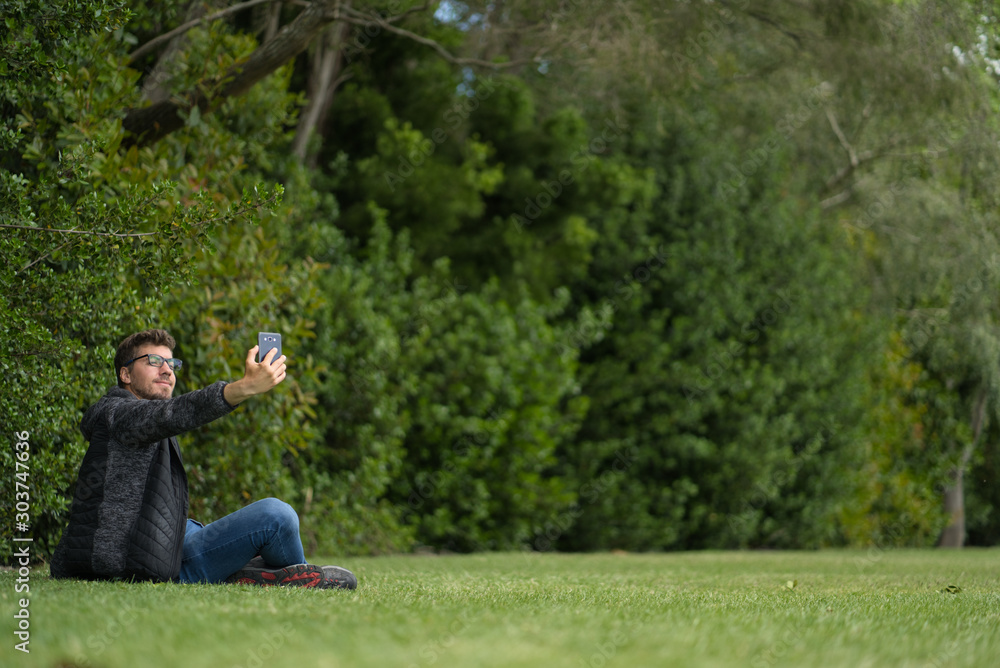 A young caucasian man wearing glasses sitting on the grass of a park and taking a selfie with his mobile