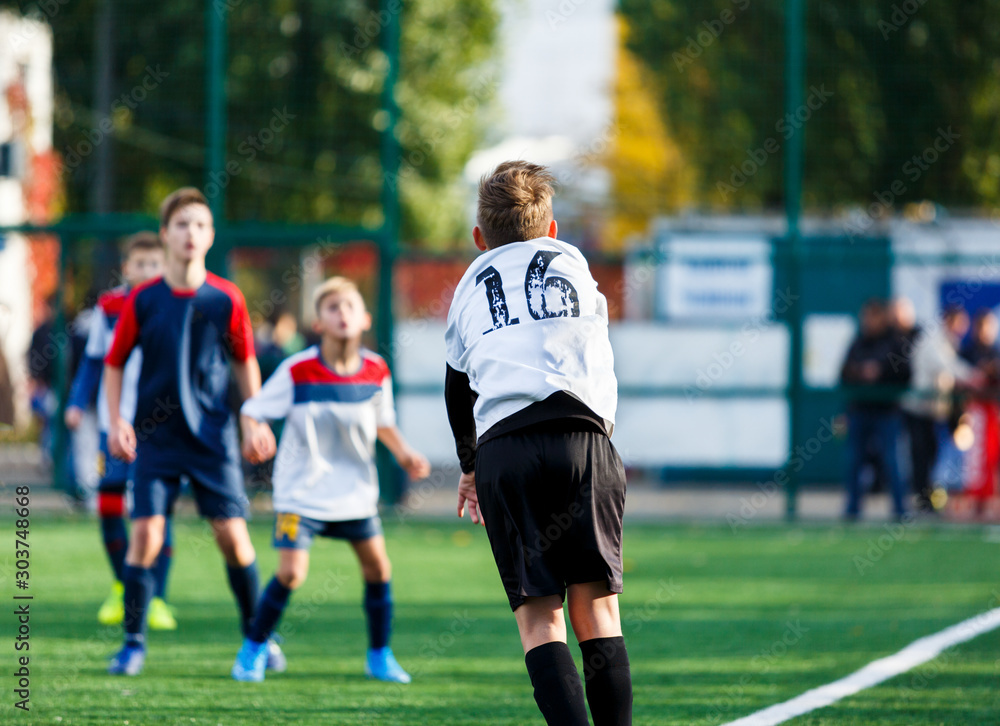 Obraz premium Boys in white and blue sportswear plays football on field, dribbles ball. Young soccer players with ball on green grass. Training, football, active lifestyle for kids concept