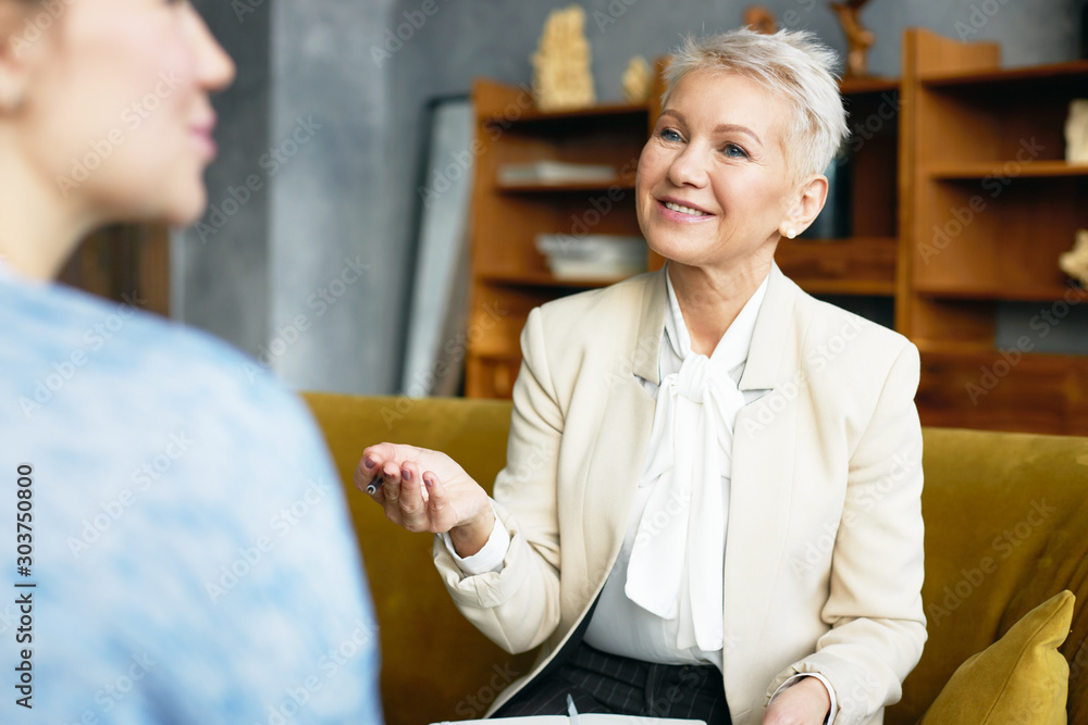 Φωτογραφία Indoor shot of positive mature female psychologist in elegant suit sitting on couch and making notes while having therapy with unrecognizable woman patient