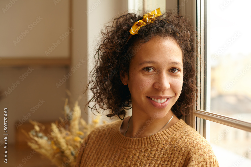 Φωτογραφία Indoor shot of charming friendly young casually dressed Latin woman with curly hair and brown skin posing by window in cozy kitchen interior, looking at camera with smile