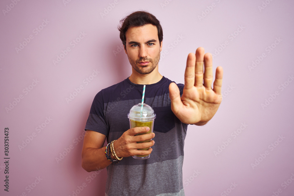 Young handsome man with tattoo drinking smoothie standing over isolated pink background with open hand doing stop sign with serious and confident expression, defense gesture