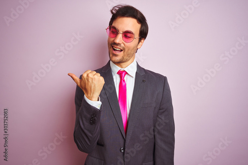 Young handsome businessman wearing suit and sunglasses over isolated pink background smiling with happy face looking and pointing to the side with thumb up.