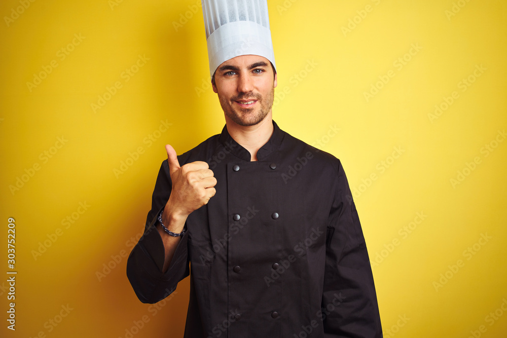 Young chef man wearing uniform and hat standing over isolated yellow background doing happy thumbs up gesture with hand. Approving expression looking at the camera showing success. Young chef man wearing uniform and hat standing over isolated yellow background doing happy thumbs up gesture with hand. Approving expression looking at the camera showing success.