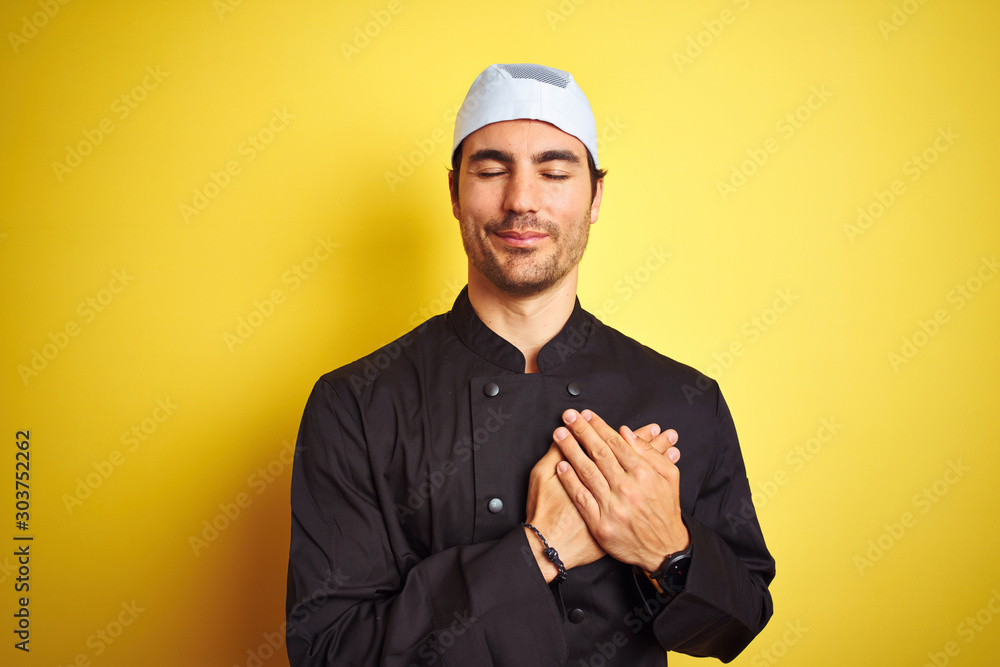 Young handsome chef man cooking wearing uniform and hat over isolated yellow background smiling with hands on chest with closed eyes and grateful gesture on face. Health concept. Young handsome chef man cooking wearing uniform and hat over isolated yellow background smiling with hands on chest with closed eyes and grateful gesture on face. Health concept.
