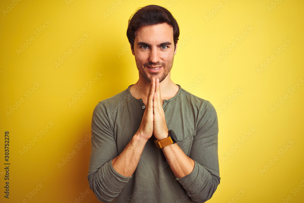 Young handsome man wearing casual t-shirt standing over isolated yellow background praying with hands together asking for forgiveness smiling confident. Young handsome man wearing casual t-shirt standing over isolated yellow background praying with hands together asking for forgiveness smiling confident.