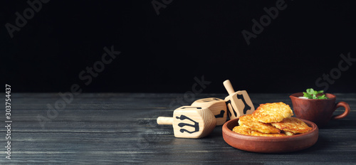 Dreidels and potato pancakes for Hanukkah on table against dark background with space for text photo