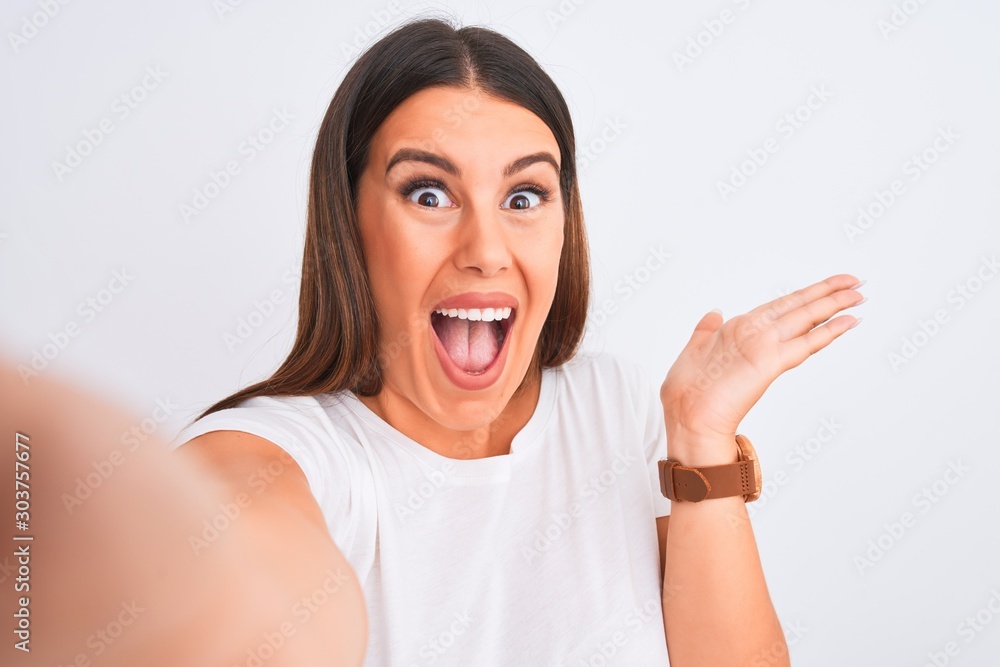Beautiful young woman taking a selfie photo using smartphone over white background very happy and excited, winner expression celebrating victory screaming with big smile and raised hands Beautiful young woman taking a selfie photo using smartphone over white background very happy and excited, winner expression celebrating victory screaming with big smile and raised hands