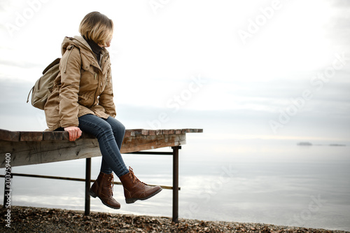 young girl in a warm jacket sits on the pier with his legs dangling and look towards the sea
