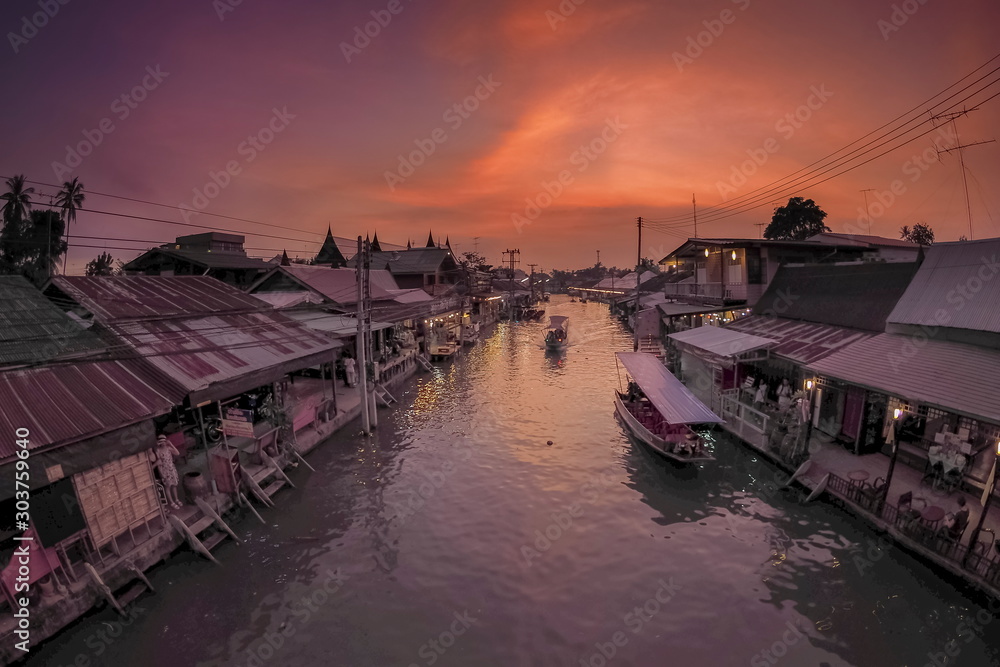 view evening of long-tail boats running in Amphawa Canal around with wooden houses and red cloudy sky background, sunset at Amphawa Floating Market, Samut Songkhram, Thailand.