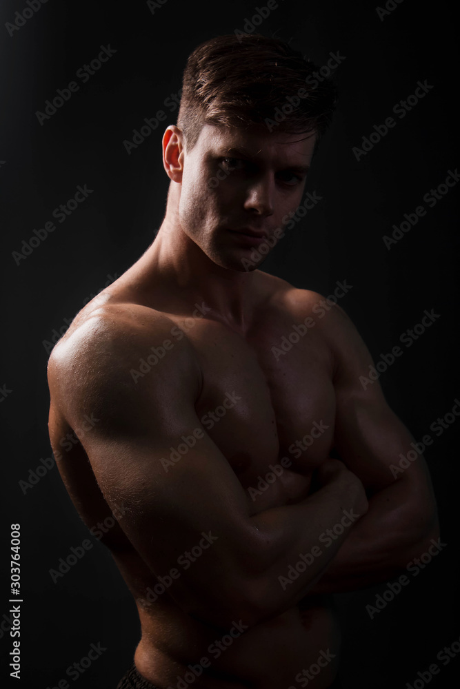 Muscular bodybuilder posing with a naked torso on a black background in the dark with backlight. Sexy man. Muscular bodybuilder posing with a naked torso on a black background in the dark with backlight. Sexy man.