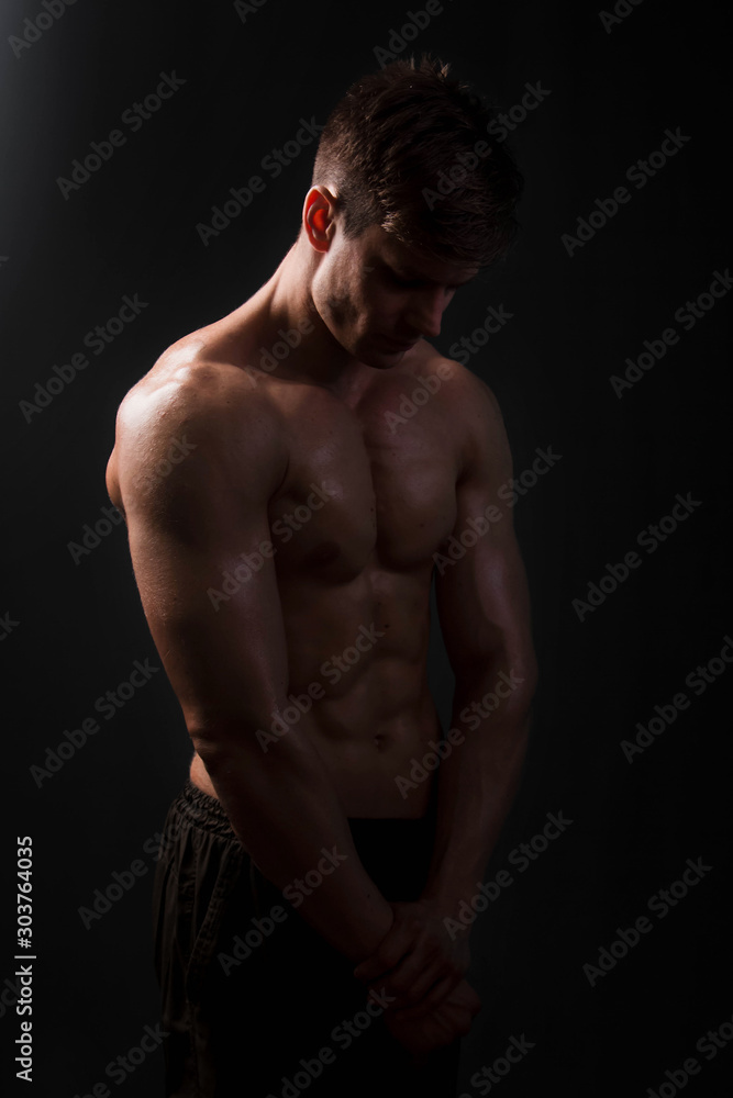 Naklejka premium Muscular bodybuilder posing with a naked torso on a black background in the dark with backlight. Sexy man.