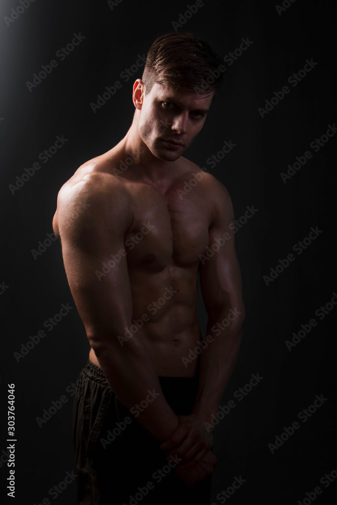 Muscular bodybuilder posing with a naked torso on a black background in the dark with backlight. Sexy man. Muscular bodybuilder posing with a naked torso on a black background in the dark with backlight. Sexy man.