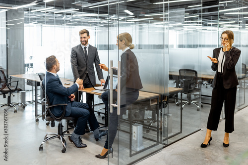 Group of strictly dressed business people having a discussion, sitting at the workplace with a computer in the modern office with glass partitions