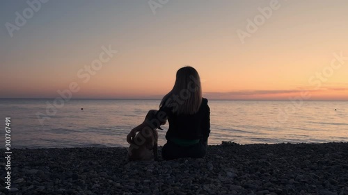 Woman with dog sitting on the beach near the sea at sunset and enjoying nature. Silhouette of a girl and her pet dog at sunset, the sea in the background.