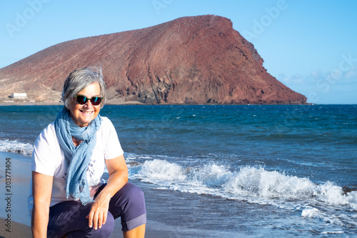 An attractive senior woman enjoys a day at the beach on a windy day. Red mountain, blue sky and sea in the background