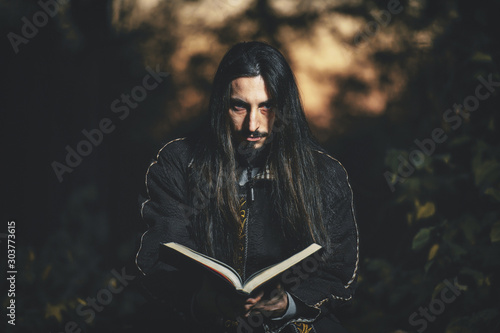 A man with long hair and beard in a black cloak in the image of a sorcerer warlock on Halloween. Dark magician the man in the background of the forest. A wizard in a dark forest with a magic book .