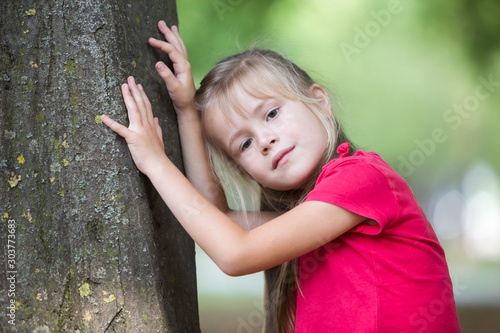 Portrait of a pretty little child girl standing near big tree trunk in summer park outdoors. © bilanol
